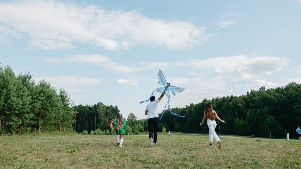 A family runs joyfully while flying a kite in a sunny park setting.
